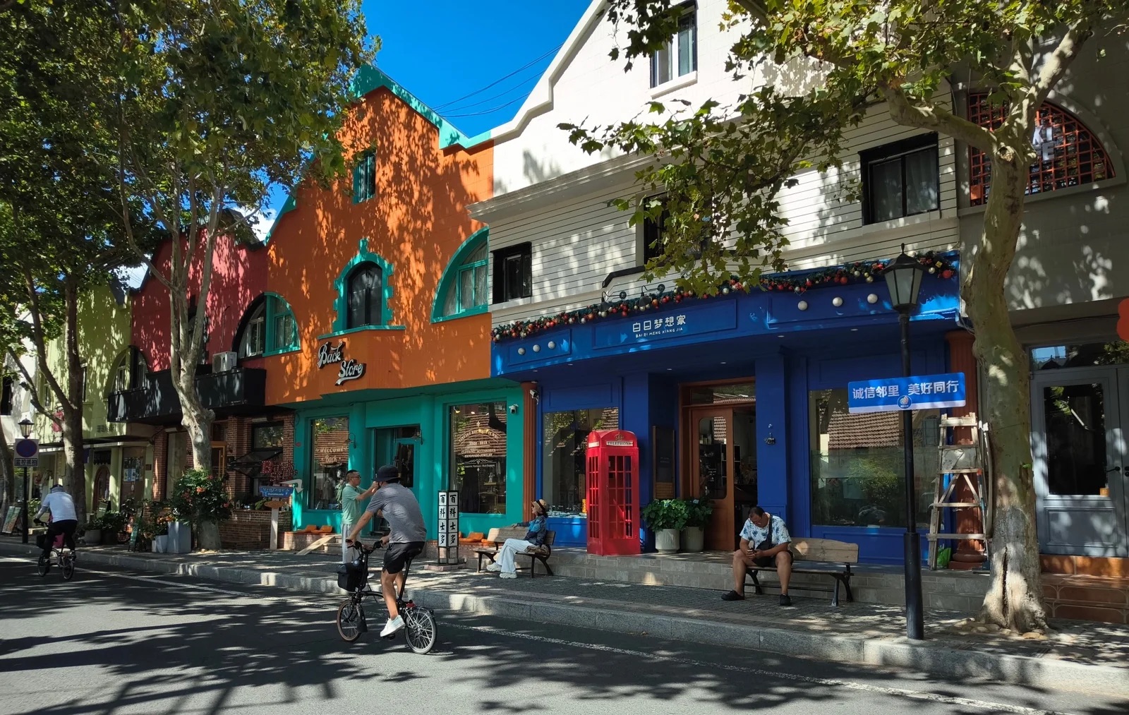 新威附路街景,橙色与蓝色的彩色小屋沿街排开,门口立着红色电话亭,梧桐树影洒满路面